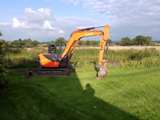 Orange excavator on grass near a wooded area under a partly cloudy sky.