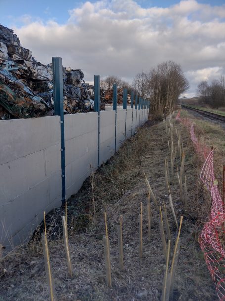 Fenced area alongside a path, with stacked materials and young plants growing.